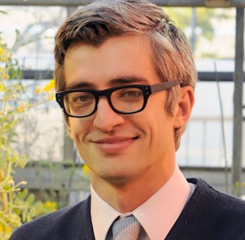 a grey haired man in glasses smirking in front of some plants 
