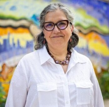 A woman in glasses and a white button up shirt in front of a colorful mural 