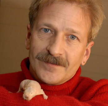 a man with a mustache in a red turtleneck holding a naked mole rat 